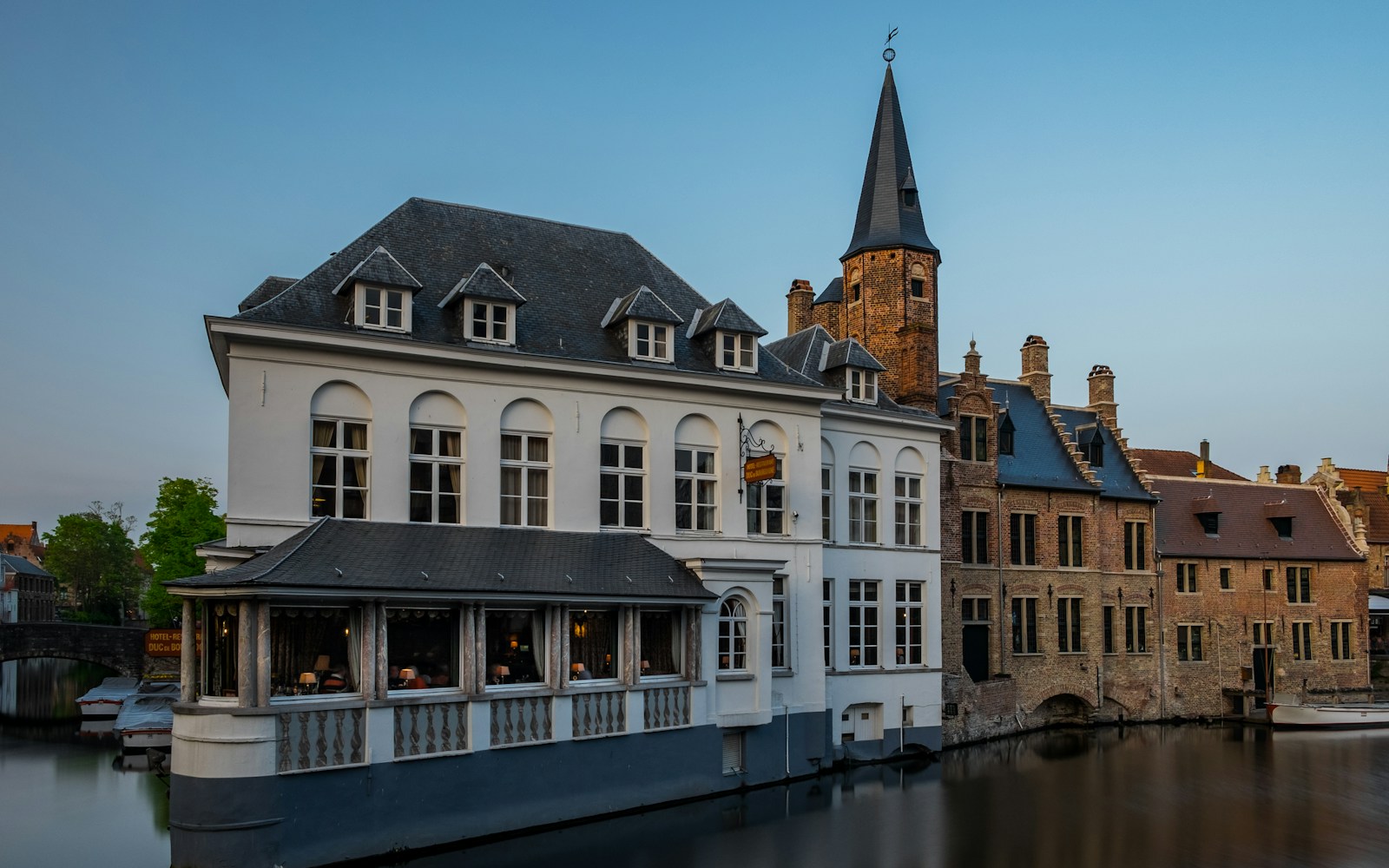 Historic step-gabled houses and a slate spire reflected in a Bruges canal at dusk
