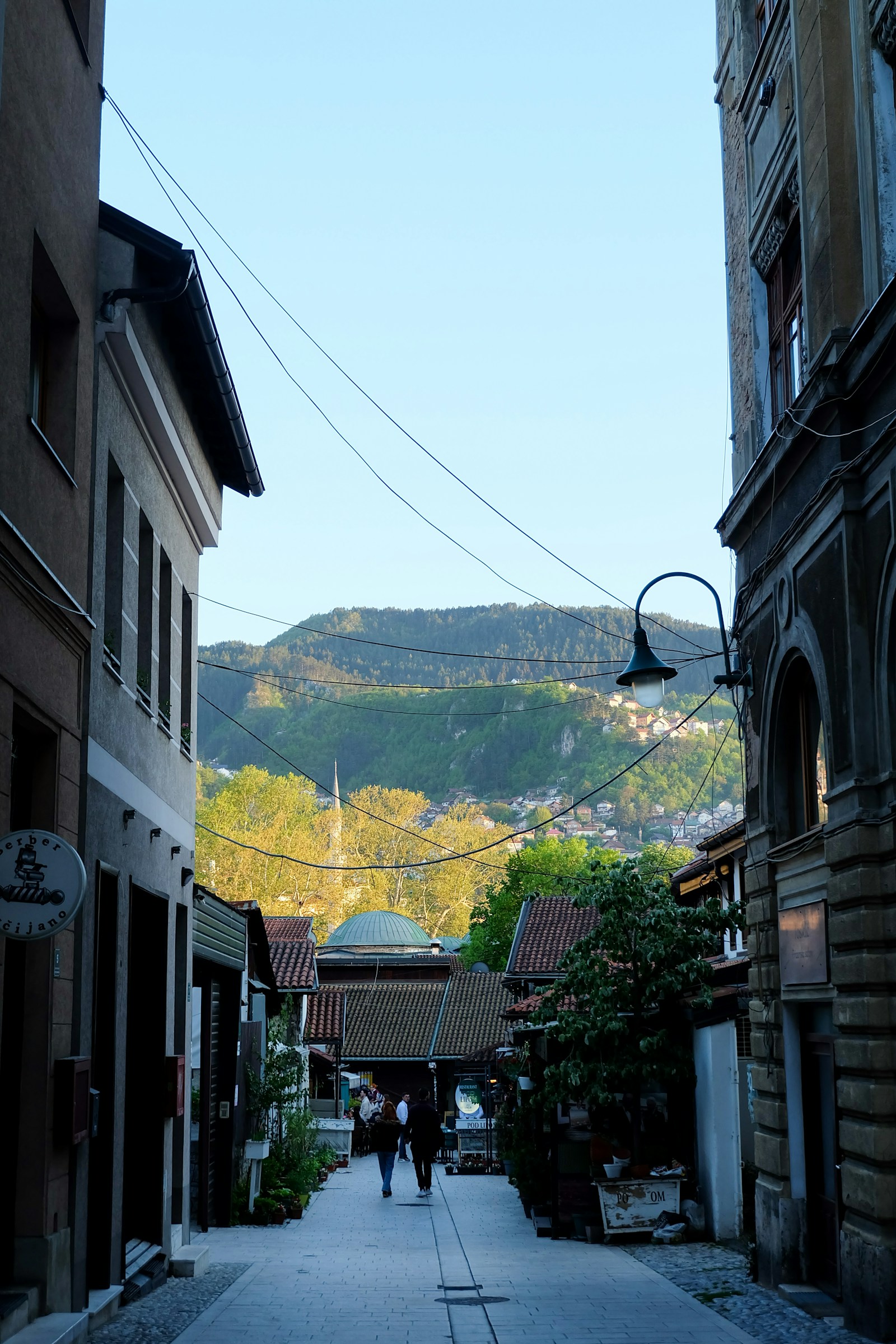 A cobbled old-town street in Sarajevo with a mosque dome and the surrounding hills behind