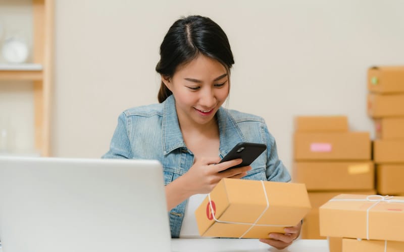 A smiling online seller checking a parcel with her phone next to a laptop