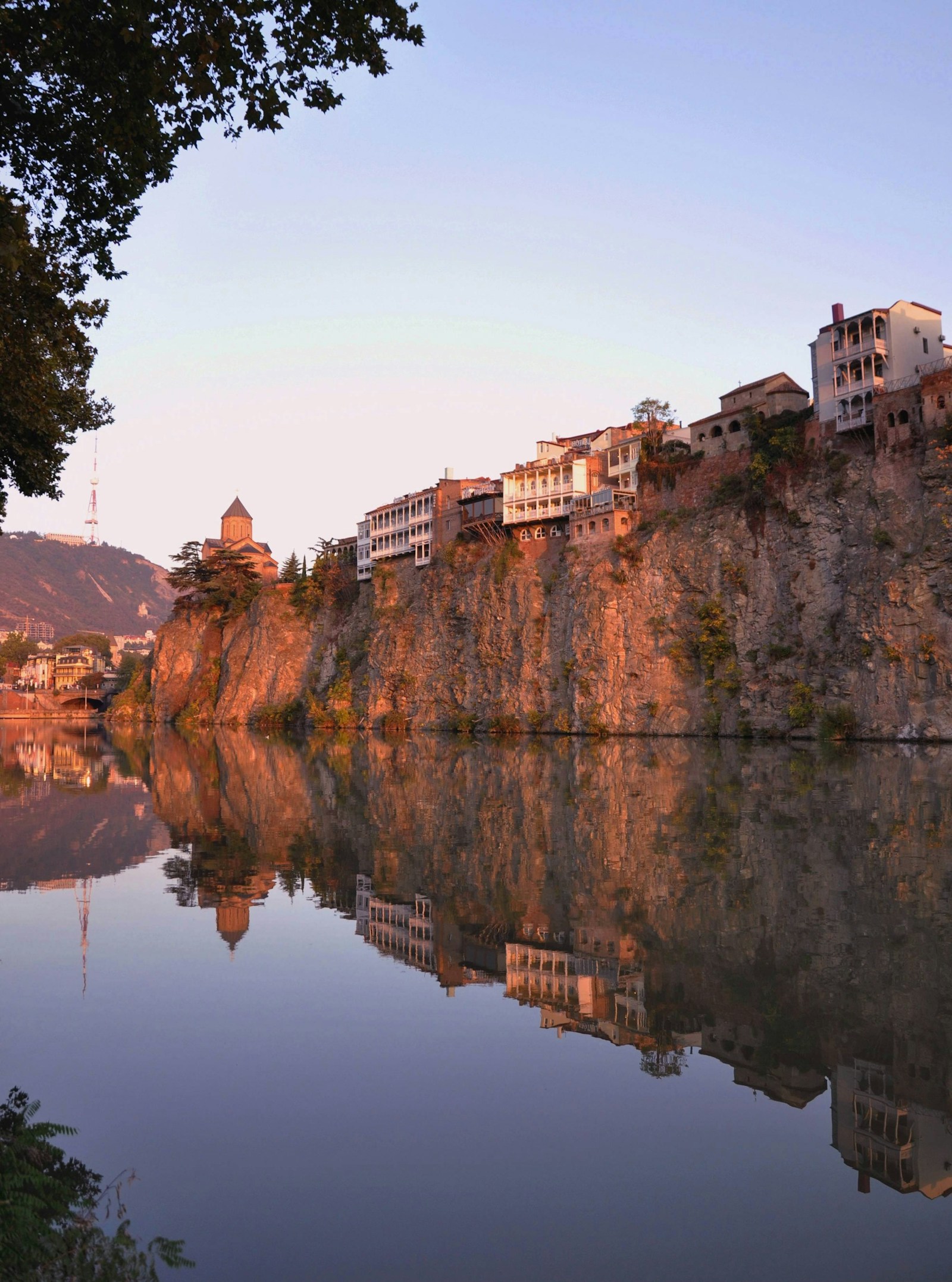 The cliffside old-town houses of Tbilisi reflected in the Mtkvari river at golden hour