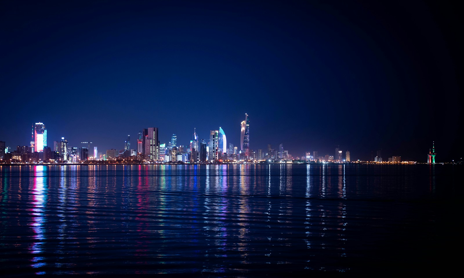 The Kuwait City skyline lit up at night across the water, with the twisting Al Hamra Tower