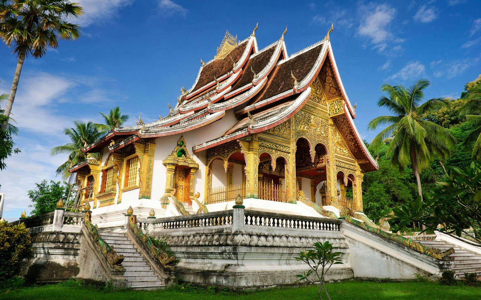 A golden Lao Buddhist temple with sweeping tiered roofs framed by palms in Luang Prabang, Laos